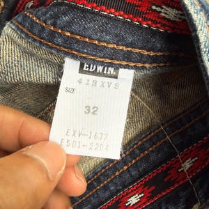 Op de afbeelding: Close-up van een denim kledinglabel. Het witte label met zwarte tekst toont het merk "EDWIN", maat informatie en het nummer "32". De denim heeft bruine stiksels en een rood-zwart patroon.