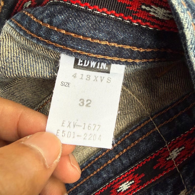 Op de afbeelding: Close-up van een denim kledinglabel. Het witte label met zwarte tekst toont het merk "EDWIN", maat informatie en het nummer "32". De denim heeft bruine stiksels en een rood-zwart patroon.