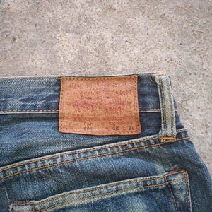May include: Close-up of blue denim jeans with a brown leather patch on the back. The patch has faded red text and the numbers 28 and 30. The jeans have visible stitching and a worn appearance.