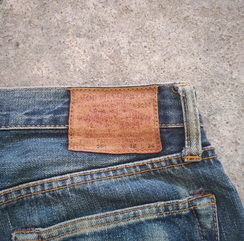 May include: Close-up of blue denim jeans with a brown leather patch on the back. The patch has faded red text and the numbers 28 and 30. The jeans have visible stitching and a worn appearance.