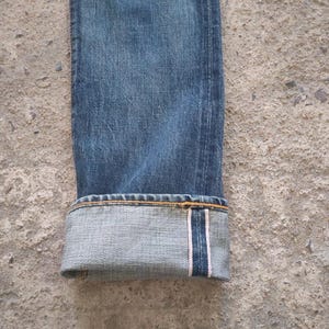 May include: Close-up of the folded cuff of blue denim jeans. The jeans have a classic, worn look with visible texture and subtle fading. The cuff reveals a lighter shade of denim with a white and red selvedge detail.