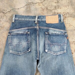 May include: Faded blue denim jeans with distressed details. The back view showcases two pockets with decorative stitching and a leather patch. The jeans have a classic five-pocket design and a button and zipper closure.