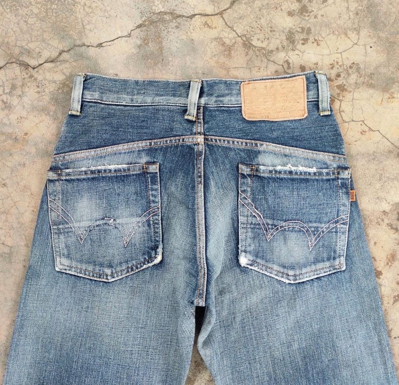 May include: Faded blue denim jeans with distressed details. The back view showcases two pockets with decorative stitching and a leather patch. The jeans have a classic five-pocket design and a button and zipper closure.