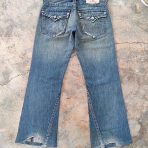 May include: Faded blue denim jeans with a distressed appearance. Features include two back pockets with decorative stitching, a leather patch, and a split hem design. The jeans have a classic five-pocket style.