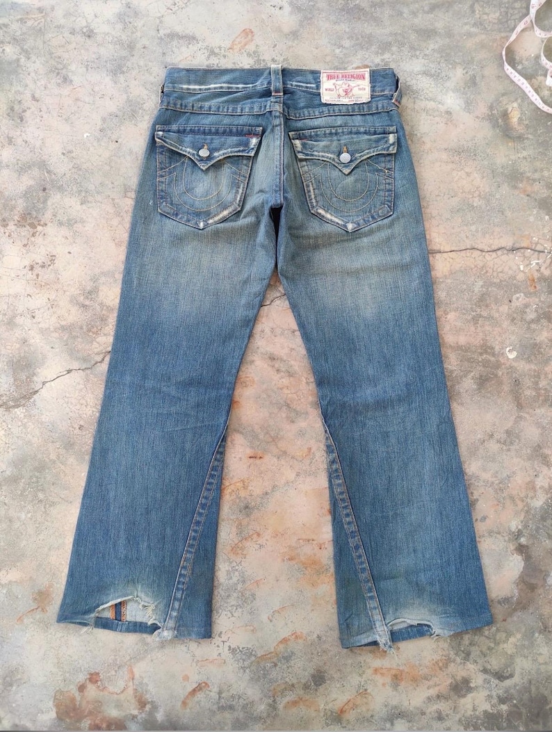 May include: Faded blue denim jeans with a distressed appearance. Features include two back pockets with decorative stitching, a leather patch, and a split hem design. The jeans have a classic five-pocket style.