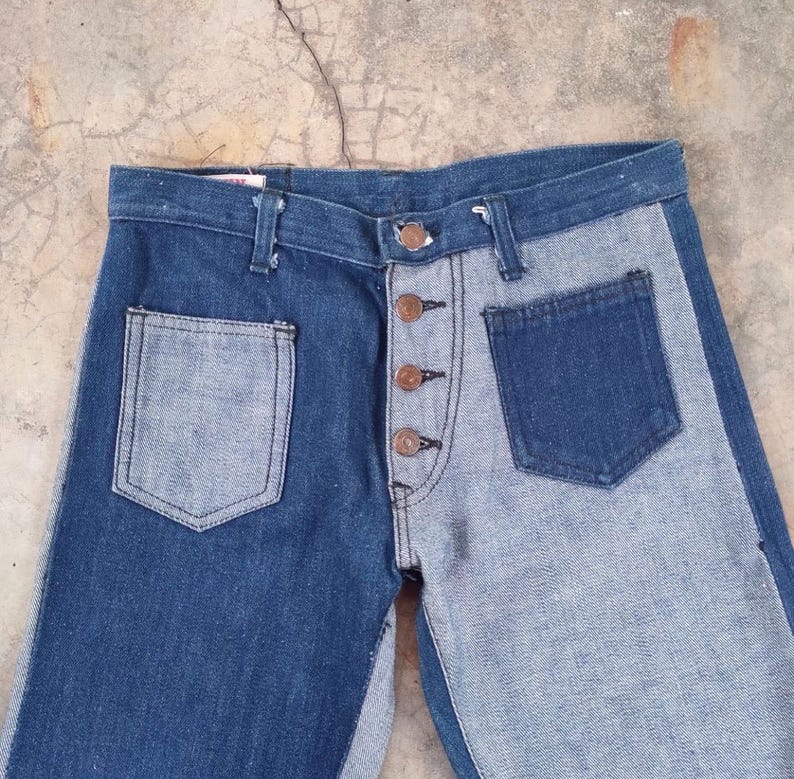 May include: Two-tone denim jeans with a button-up fly and two front pockets. The jeans feature a contrasting design, with one side in a darker blue and the other in a lighter blue wash.