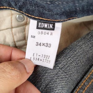 Puede incluir: Primer plano de una etiqueta de jeans de mezclilla con la marca "EDWIN". La etiqueta muestra la talla "34x33". Los jeans son de color azul cl&aacute;sico, con costuras visibles y un bot&oacute;n. La etiqueta tambi&eacute;n incluye el n&uacute;mero de modelo "0504 R".