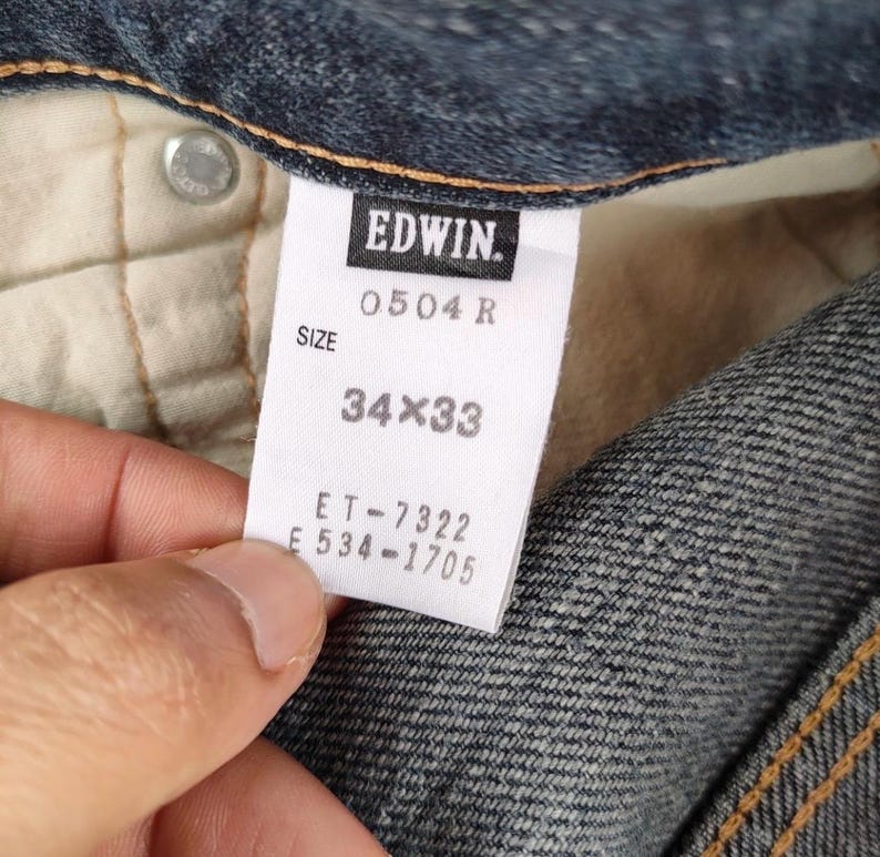 Puede incluir: Primer plano de una etiqueta de jeans de mezclilla con la marca "EDWIN". La etiqueta muestra la talla "34x33". Los jeans son de color azul cl&aacute;sico, con costuras visibles y un bot&oacute;n. La etiqueta tambi&eacute;n incluye el n&uacute;mero de modelo "0504 R".