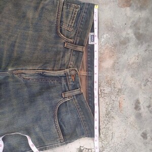 May include: Faded blue denim jeans with a classic five-pocket design. A measuring tape shows the length of the jeans. The jeans have a button and zipper closure and belt loops. The brand label is visible.