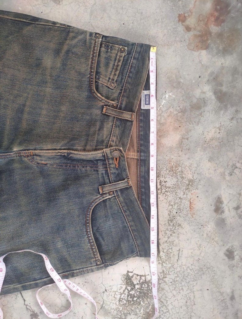 May include: Faded blue denim jeans with a classic five-pocket design. A measuring tape shows the length of the jeans. The jeans have a button and zipper closure and belt loops. The brand label is visible.