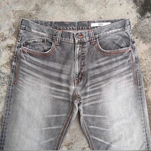 K&ouml;nnte beinhalten: Graue Jeans mit orangefarbenen N&auml;hten. Die Jeans haben ein klassisches F&uuml;nf-Taschen-Design, einen Knopf- und Rei&szlig;verschluss und den Markennamen "XU-S" auf dem Etikett. Die Jeans haben einen verwaschenen Effekt.