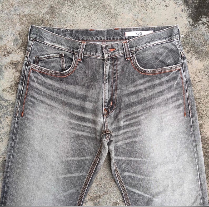 K&ouml;nnte beinhalten: Graue Jeans mit orangefarbenen N&auml;hten. Die Jeans haben ein klassisches F&uuml;nf-Taschen-Design, einen Knopf- und Rei&szlig;verschluss und den Markennamen "XU-S" auf dem Etikett. Die Jeans haben einen verwaschenen Effekt.