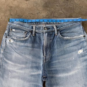 Pu&ograve; includere: Jeans in denim blu chiaro con chiusura a bottone e dettagli strappati. La vita misura circa 43 cm di larghezza. Presenta un classico stile a cinque tasche e un lavaggio sbiadito.