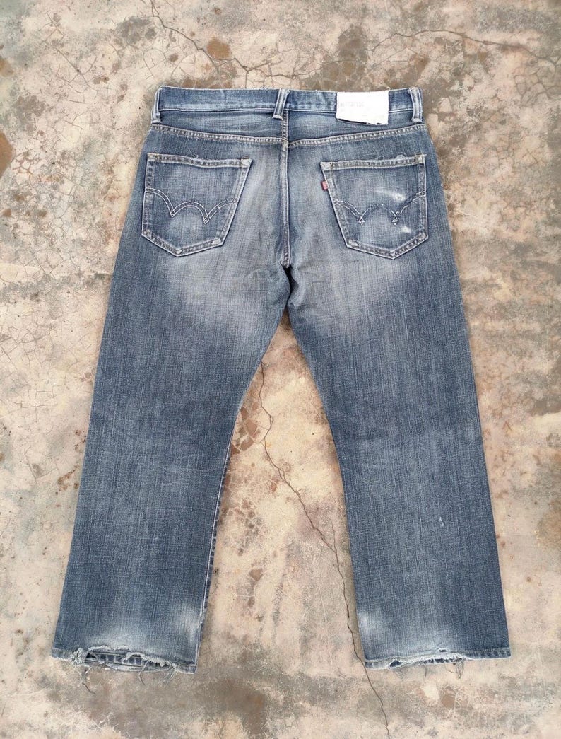 Puede incluir: Vaqueros de mezclilla azul descolorido con un dise&ntilde;o cl&aacute;sico de cinco bolsillos. Los vaqueros tienen un aspecto desgastado con detalles desgastados en los bolsillos traseros y los dobladillos. Una peque&ntilde;a etiqueta roja es visible en uno de los bolsillos traseros.