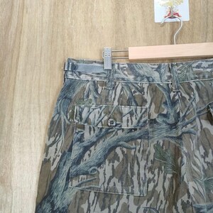 May include: Camouflage trousers with a buttoned pocket and belt loops. The trousers feature a pattern of brown, green, and grey tree-like designs. The trousers are hanging on a wooden hanger.