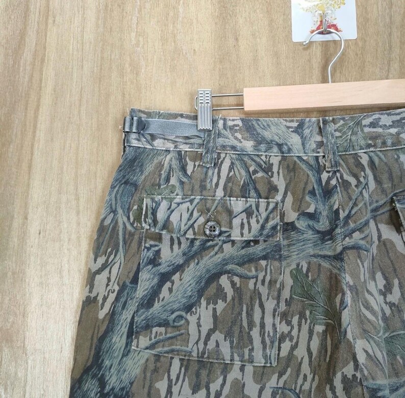 May include: Camouflage trousers with a buttoned pocket and belt loops. The trousers feature a pattern of brown, green, and grey tree-like designs. The trousers are hanging on a wooden hanger.