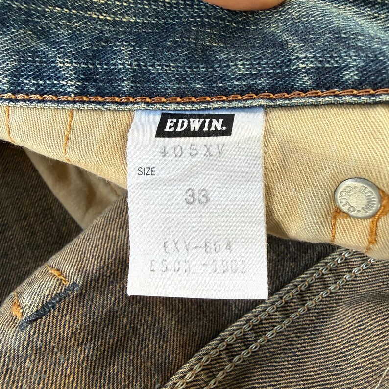 Puede incluir: Primer plano de una etiqueta de jeans de mezclilla con la marca "EDWIN" en un rect&aacute;ngulo negro. La etiqueta muestra la talla "33" y otros detalles. Los jeans son azules con un aspecto desgastado.