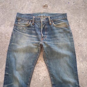 May include: Faded blue denim jeans with a classic five-pocket design. The jeans show signs of wear and fading, with a button fly and belt loops. The jeans are laid flat on a concrete surface.