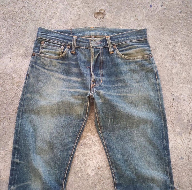 May include: Faded blue denim jeans with a classic five-pocket design. The jeans show signs of wear and fading, with a button fly and belt loops. The jeans are laid flat on a concrete surface.