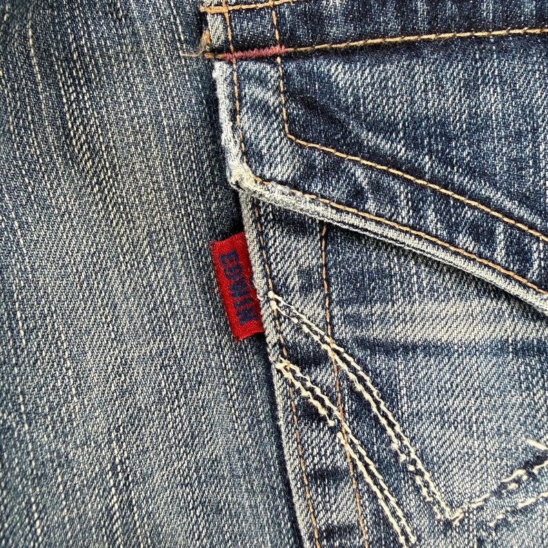 May include: Close-up of a section of blue denim jeans. The image highlights the back pocket with white stitching and a red Edwin label. The denim has a worn, textured appearance.
