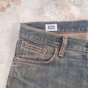 May include: Close-up of blue denim jeans with a faded wash. The jeans feature a classic five-pocket design, with visible stitching and rivets. The brand name "EDWIN INTERNATIONAL BASIC" is displayed on a small label.