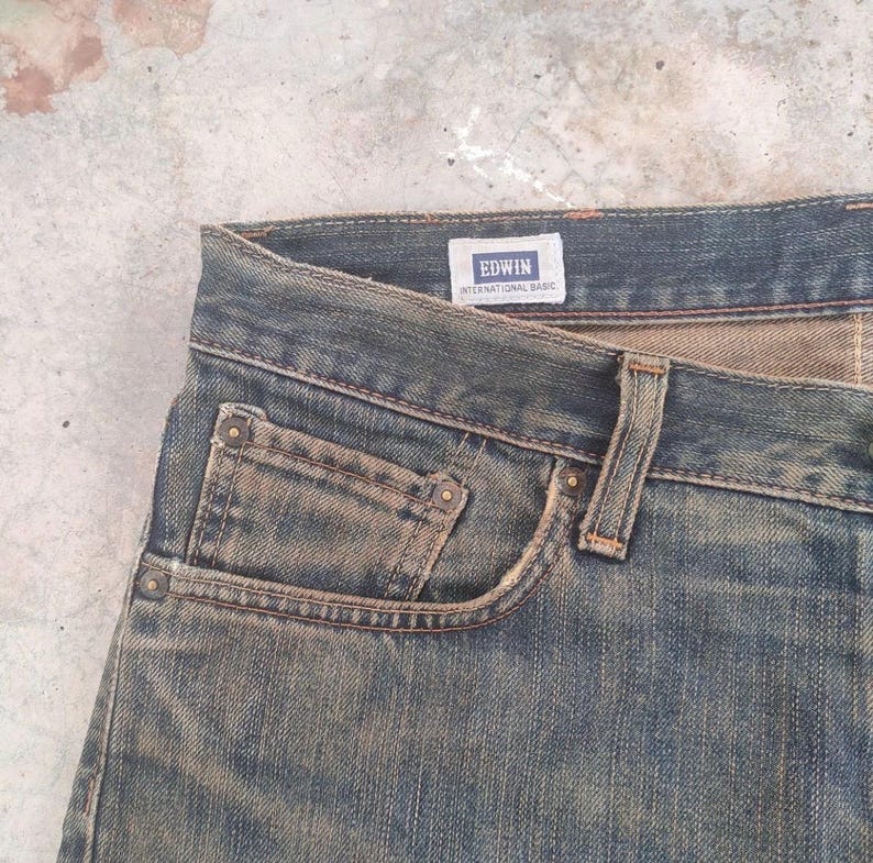 May include: Close-up of blue denim jeans with a faded wash. The jeans feature a classic five-pocket design, with visible stitching and rivets. The brand name "EDWIN INTERNATIONAL BASIC" is displayed on a small label.