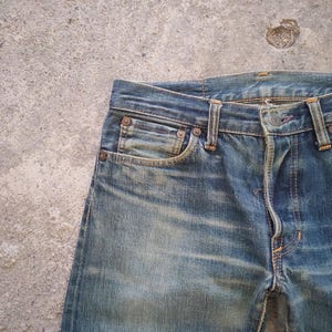 May include: Close-up of blue denim jeans with a faded wash. The jeans feature classic five-pocket styling, a button closure, and belt loops. The fabric shows signs of wear, with subtle fading and whiskering.