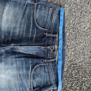 Pu&ograve; includere: Primo piano di jeans blu in denim con lavaggio sbiadito. Un metro a nastro blu mostra una lunghezza di circa 40 cm. I jeans hanno un classico stile a cinque tasche e una chiusura con bottone.