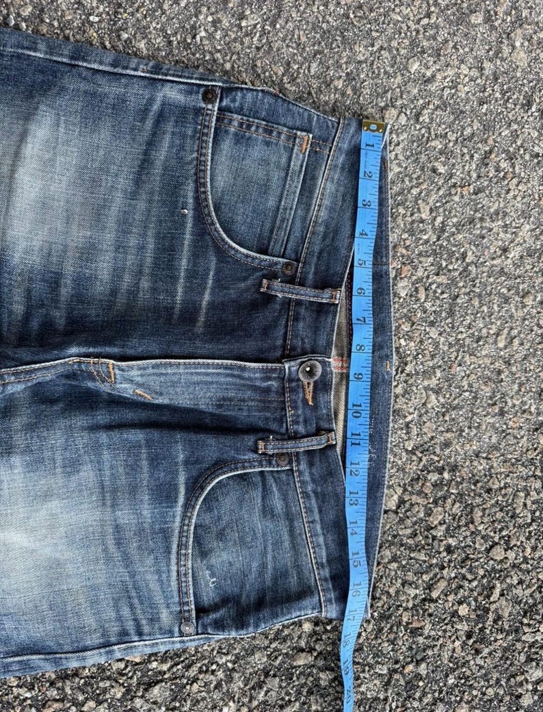 Pu&ograve; includere: Primo piano di jeans blu in denim con lavaggio sbiadito. Un metro a nastro blu mostra una lunghezza di circa 40 cm. I jeans hanno un classico stile a cinque tasche e una chiusura con bottone.