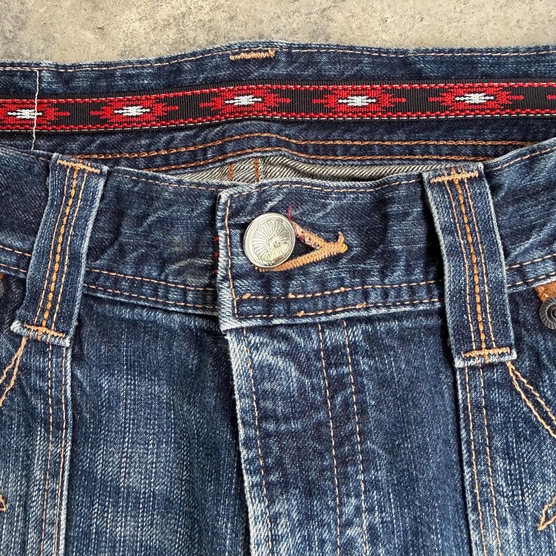 Op de afbeelding: Close-up van een blauwe spijkerbroek met oranje stiksels. De tailleband heeft een decoratief zwart-rood patroonlint. Een zilveren knoop met een zonnesterontwerp sluit de gulp. De jeans vertoont tekenen van slijtage.