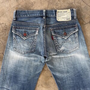 May include: Faded blue denim jeans with unique pocket designs and red accents. The back pockets feature a distinctive embroidered pattern. A label on the waistband reads "BLUE TRIP" and "503".