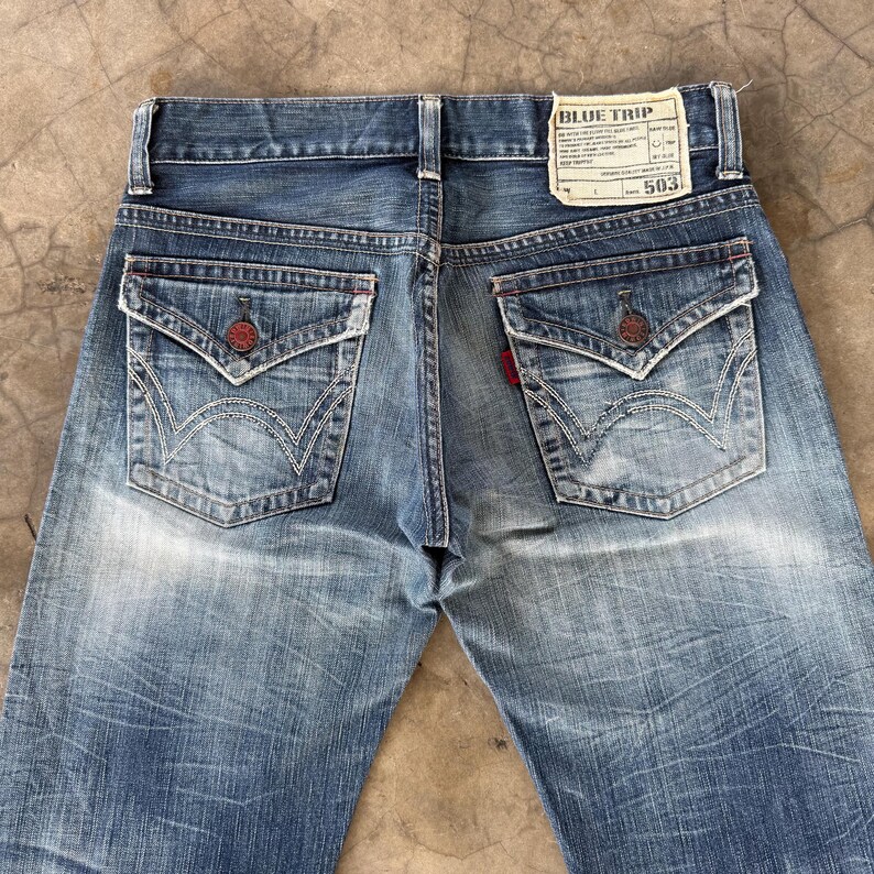 May include: Faded blue denim jeans with unique pocket designs and red accents. The back pockets feature a distinctive embroidered pattern. A label on the waistband reads "BLUE TRIP" and "503".