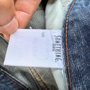 May include: Close-up of blue denim jeans with a white tag. The tag reads "SOMETHING." and "EDWIN." in black text. The word "SIZE" is also visible on the tag. The jeans have a classic five-pocket design.