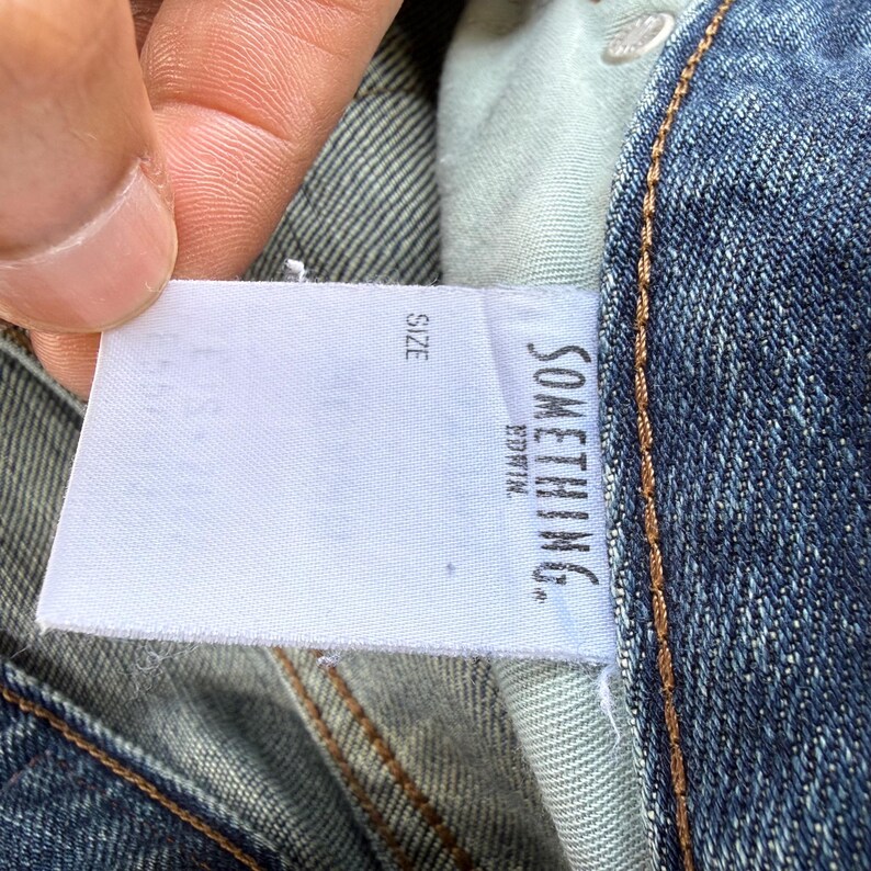 May include: Close-up of blue denim jeans with a white tag. The tag reads "SOMETHING." and "EDWIN." in black text. The word "SIZE" is also visible on the tag. The jeans have a classic five-pocket design.