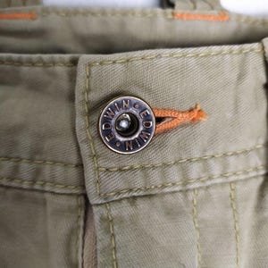 May include: Close-up of khaki-colored pants with a metal button. The button is silver with the word "EDWIN" around the edge. Orange stitching is visible near the button and along the seams of the pants.