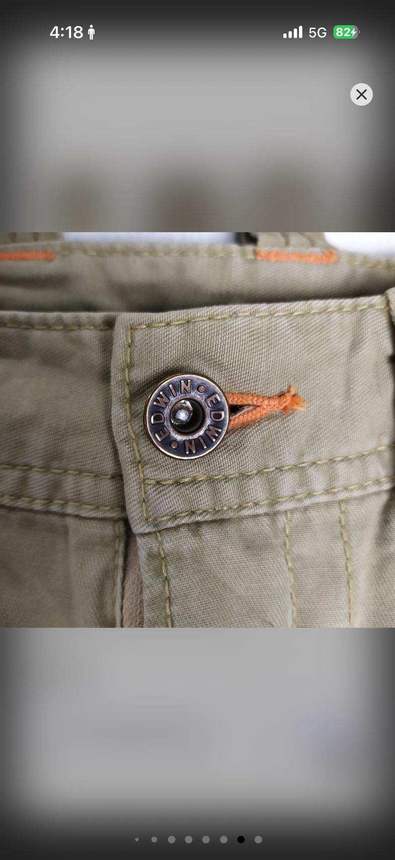 May include: Close-up of khaki-colored pants with a metal button. The button is silver with the word "EDWIN" around the edge. Orange stitching is visible near the button and along the seams of the pants.