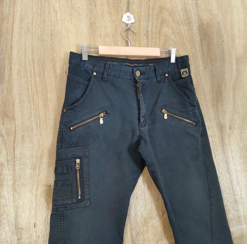 Pu&ograve; includere: Pantaloni cargo blu navy scuro con cerniere color oro sulle tasche e sulla patta. I pantaloni sono appesi a una gruccia di legno su uno sfondo chiaro a venature di legno. Stile classico e casual.