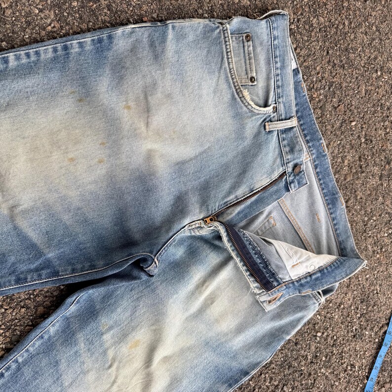 May include: Light blue denim jeans with a partially unzipped fly. The jeans show signs of wear with faded areas and some light stains. The classic five-pocket design is visible, and the jeans are laid out on a textured surface.