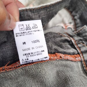 K&ouml;nnte beinhalten: Ein wei&szlig;es Etikett mit japanischem Text und Pflegesymbolen ist an einem grauen Denimstoff befestigt. Das Etikett lautet "100% Baumwolle" und "Made in China". Das Etikett enth&auml;lt auch Waschanweisungen und einen Hinweis auf Farbausblutung.