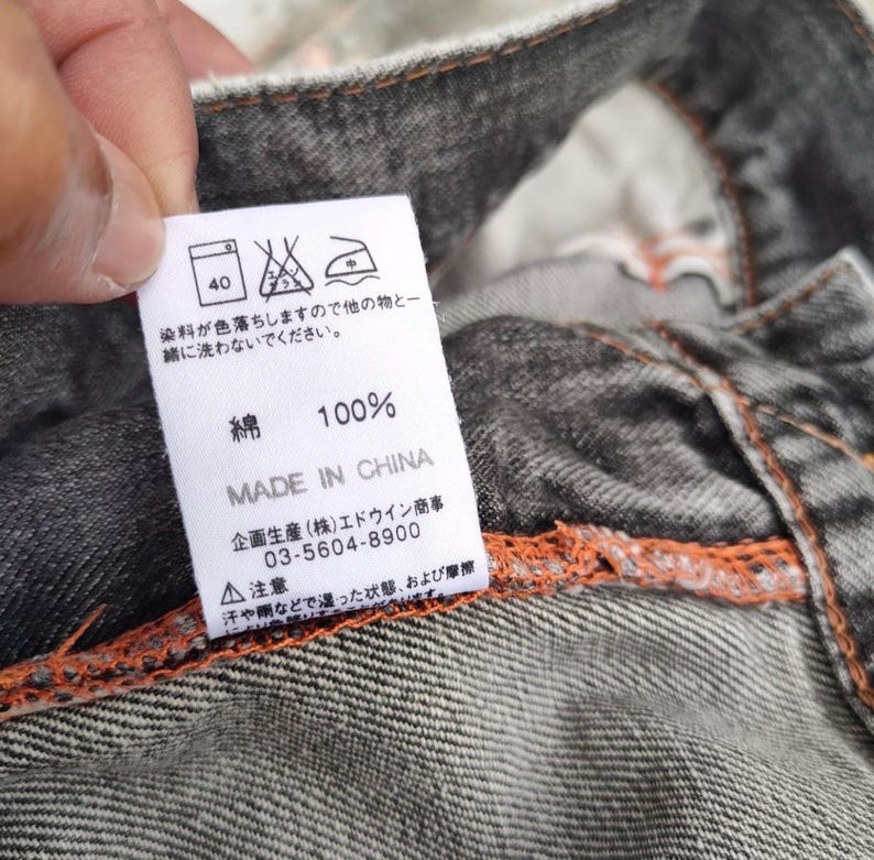K&ouml;nnte beinhalten: Ein wei&szlig;es Etikett mit japanischem Text und Pflegesymbolen ist an einem grauen Denimstoff befestigt. Das Etikett lautet "100% Baumwolle" und "Made in China". Das Etikett enth&auml;lt auch Waschanweisungen und einen Hinweis auf Farbausblutung.