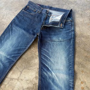 May include: A pair of blue denim jeans with a faded wash effect. The jeans are unbuttoned and unzipped, showcasing the interior details. The fabric shows wear and tear, with visible fading and lighter patches.