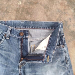 May include: Close-up of blue denim jeans with a partially unzipped fly. The jeans have a button closure and a classic five-pocket design. The fabric shows signs of wear, with a faded appearance.