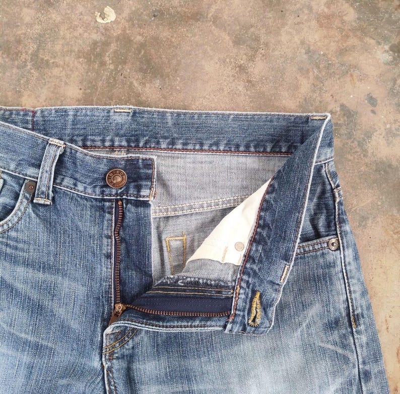 May include: Close-up of blue denim jeans with a partially unzipped fly. The jeans have a button closure and a classic five-pocket design. The fabric shows signs of wear, with a faded appearance.