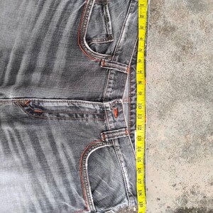 K&ouml;nnte beinhalten: Graue Jeans mit orangefarbenen N&auml;hten. Die Jeans haben ein klassisches F&uuml;nf-Taschen-Design und einen geraden Beinverlauf. Ein gelbes Ma&szlig;band zeigt die L&auml;nge der Jeans, ca. 51 cm.
