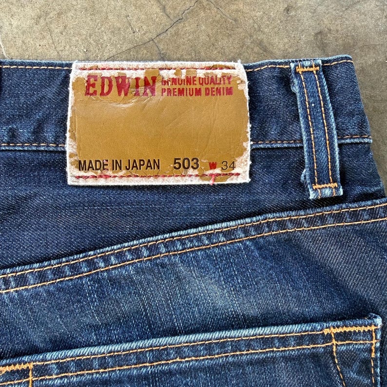 May include: Close-up of dark blue denim jeans with visible stitching and a brown leather patch. The patch reads "EDWIN" and "GENUINE QUALITY PREMIUM DENIM", with "MADE IN JAPAN 503 W 34" below.