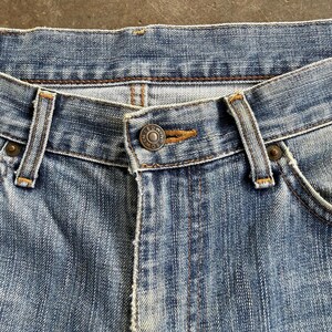 May include: Close-up of light blue denim jeans. The jeans feature a button closure with the brand name "EDWIN" on the button. The jeans have classic five-pocket styling with visible stitching and belt loops.