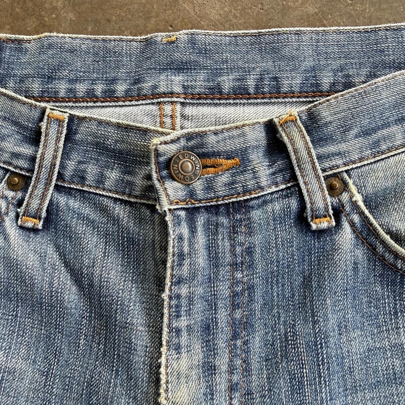 May include: Close-up of light blue denim jeans. The jeans feature a button closure with the brand name "EDWIN" on the button. The jeans have classic five-pocket styling with visible stitching and belt loops.