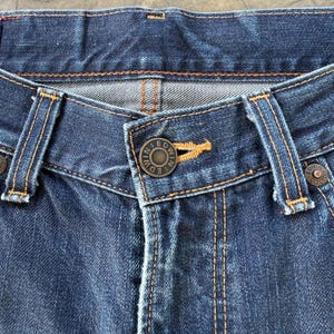 May include: Close-up of a pair of dark blue denim jeans. The jeans feature a button closure with the brand name "EDWIN" on the button. Visible are the belt loops, stitching, and rivets.