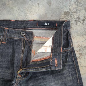 Pu&ograve; includere: Primo piano di jeans in denim lavato scuro con cuciture arancioni. I jeans hanno una chiusura con cerniera e un bottone. L'etichetta del marchio "XU-S" &egrave; visibile sulla cintura. I jeans sono leggermente sbiaditi.