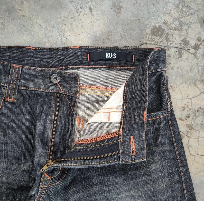 Pu&ograve; includere: Primo piano di jeans in denim lavato scuro con cuciture arancioni. I jeans hanno una chiusura con cerniera e un bottone. L'etichetta del marchio "XU-S" &egrave; visibile sulla cintura. I jeans sono leggermente sbiaditi.